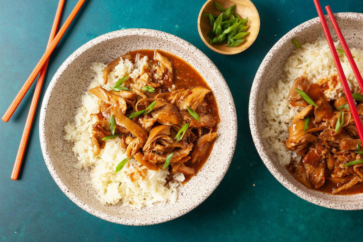 Overhead shot of two speckled bowls filled with white rice topped with tender shredded honey-garlic chicken in a glossy brown sauce, garnished with sliced green onions; chopsticks rest beside the bowls on a teal surface;