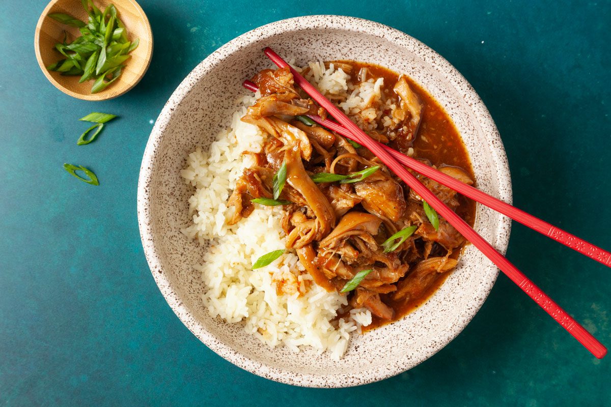 Overhead shot of two speckled bowls filled with white rice topped with tender shredded honey-garlic chicken in a glossy brown sauce, garnished with sliced green onions; chopsticks rest beside the bowls on a teal surface;