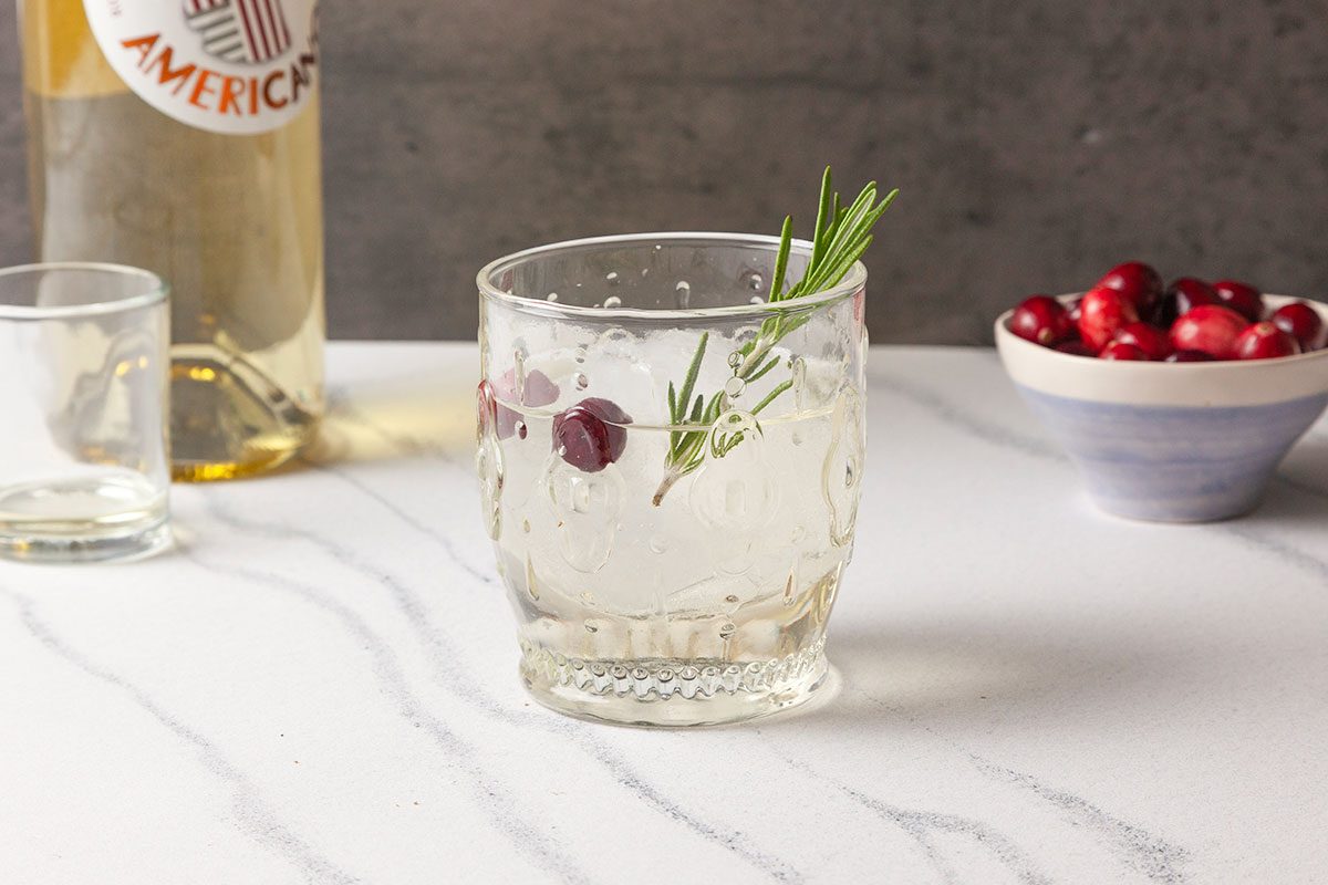 Front-facing shot of a Snowy Negroni served over ice in a rocks glass, garnished with a rosemary sprig and floating cranberries on a marble surface.