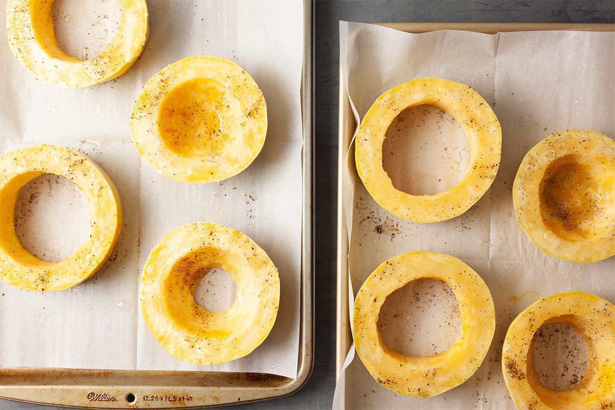 Overhead shot of two parchment-lined baking sheets hold hollowed rings of squash arranged in neat rows; Each ring is lightly coated with oil and sprinkled with seasoning, showing a glossy, golden surface;