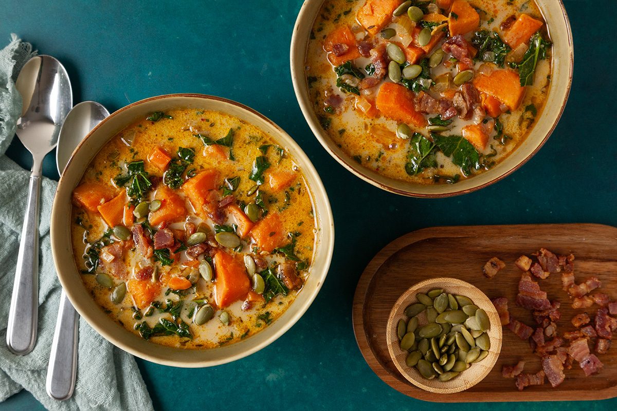 Overhead shot of two bowls of Easy Sweet Potato Chowder filled with tender sweet potato chunks, leafy greens, crispy bacon, and pumpkin seeds in a creamy, golden broth; A wooden board with chopped bacon and a small bowl of pumpkin seeds sits beside the bowls on a dark teal surface;