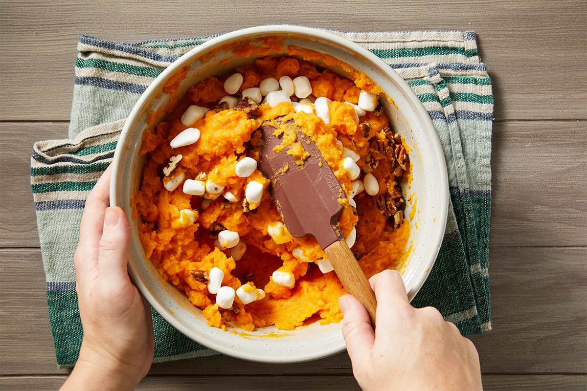 Overhead shot of a person mixing mashed sweet potatoes and mini marshmallows in a bowl with a spatula, set on a striped kitchen towel atop a wooden surface;