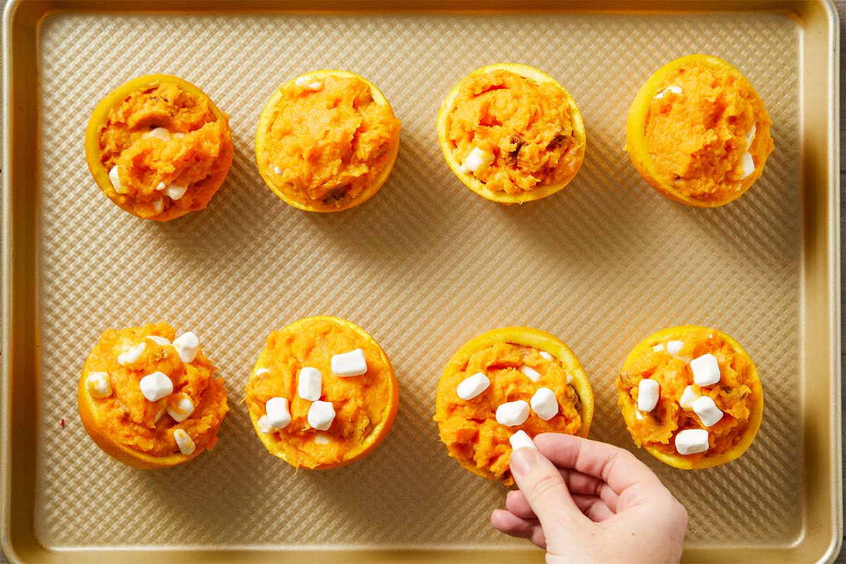 Overhead shot of a hand topping mini sweet potato casseroles in yellow cups with marshmallows on a baking sheet; four casseroles are already topped, and four remain plain;
