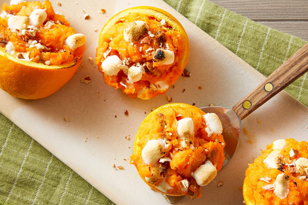 Overhead shot of Sweet Potato Orange Cups on a platter, with one cup lifted by a serving spatula; The toasted marshmallows and chopped nuts are clearly visible, set against a green striped kitchen towel;
